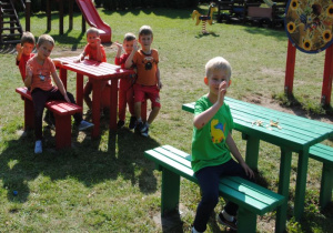 dzieci siedzą i stoją obok ławek i stolików w ogrodzie wg ich kolorów i ubrań (czerwony i zielony)